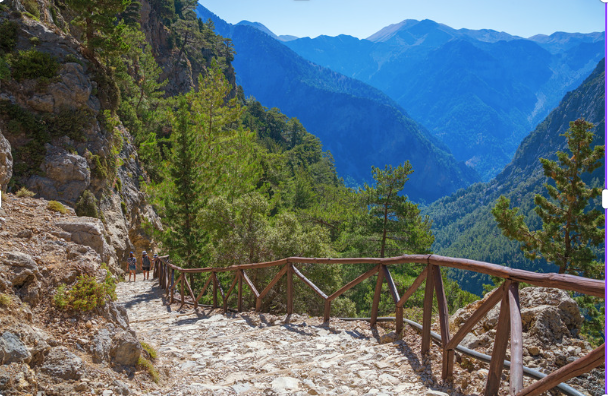 Scenic view of the Samaria Gorge in Crete, with a stone hiking trail and wooden railings winding through a dramatic mountainous landscape covered in lush greenery.